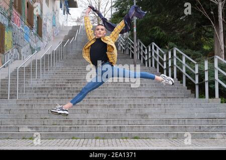 Hübscher junger Mann, der Make-up und Kopfhörer trägt und einen Ballettsprung auf einer Straßentreppe macht. Nicht binäre androgyne Kerl. Stockfoto