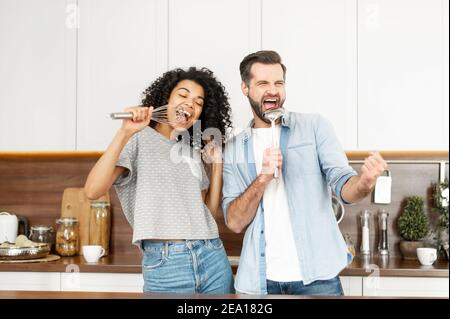 Merry multi-ethnischen Paar hat einen Spaß, Narr rund um die Vorbereitung Abendessen zu Hause. Eine Afrikanerin und ein Kaukasier tanzen in der Küche und singen hinein Stockfoto