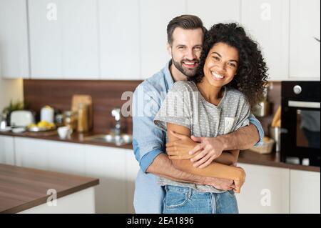 Süßes multiethnisches Paar in der Liebe steht in Umarmungen in der Küche. Heiter eine afrikanische Frau und ein Kaukasusmann schauen auf die Kamera und lächeln toothy Stockfoto