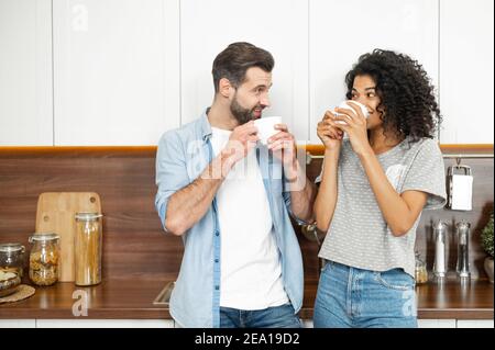Interracial paar trinkt morgens Kaffee in der Küche zu Hause. Eine Afrikanerin und ein Kaukasianer genießen zu Hause heiße Getränke Stockfoto
