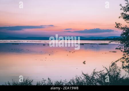 Lange Belichtung Vanille bunten Himmel und See Reflexion mit Bergen im Hintergrund. Verschwommenes wildes Gras weht im Wind im Vordergrund Stockfoto