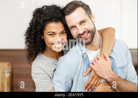 Nahaufnahme Porträt von glücklichen multi-ethnischen Paar in der Liebe. Eine Afrikanerin umarmt einen Kaukasier von hinten, sie schaut mit einem fröhlichen Blick auf die Kamera Stockfoto
