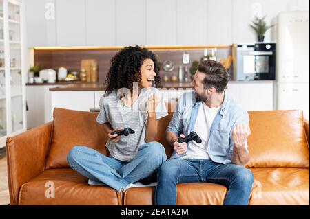 Aufgeregt multirassischen Paar spielt Videospiel, eine afrikanische Frau und ein kaukasischer Mann halten drahtlose Joysticks Blick auf einander bereit zu kämpfen Stockfoto