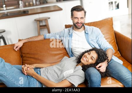 Lächelndes multiethnisches Paar in der Liebe ruht zu Hause zusammen, verbringt ein faules Wochenende auf der Couch im gemütlichen Wohnzimmer. Afrikanische Frau liegt auf den Schoß von Stockfoto