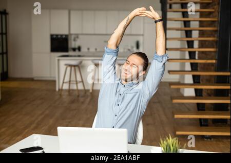 Fröhlicher junger Geschäftsmann streckt die Arme im Büro mit Laptop. Lächelnder Hipster, der sich nach erfolgreicher Arbeit am Arbeitsplatz entspannt Stockfoto