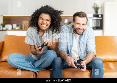 Ein multirassisches Paar genießt Zeit spielen Videospiel auf der Konsole, aufgeregt und angespannt junge afrikanische Frau und kaukasischen Kerl mit einem Joysticks versuchen, Stockfoto