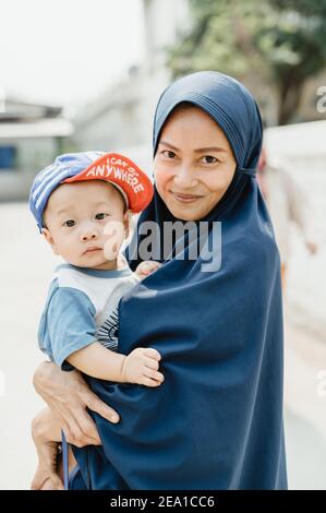 Thailändische muslimische Frau trägt einen blauen Hijab lächelt an der Kamera, hält ein Baby mit einer Mütze, schaut auf die Kamera, mit sonnigen hellen Hintergrund. Stockfoto