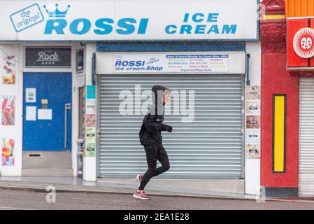 Southend on Sea, Essex, Großbritannien. Februar 2021, 7th. Storm Darcy erreichte Southend on Sea später als ursprünglich prognostiziert, wobei das Gebiet über Nacht nass blieb. Schnee und Schneestürmchen kamen um 7am an, was Jogger nicht abschreckte. COVID 19-Sperrwarnungen bleiben erhalten. Ein Rüde in Leggings und Kapuzenpulli läuft an geschlossenen Unternehmen am Meer vorbei. Geschlossen Rossi Eisdiele Stockfoto