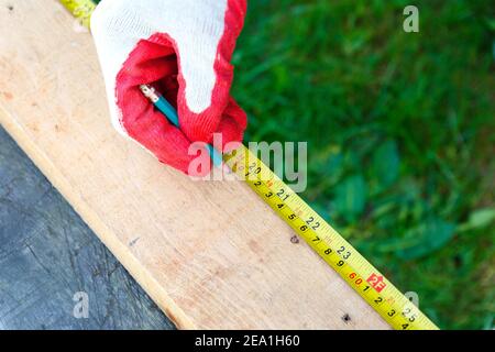 Eine Männerhand in einem Arbeitshandschuh mit einem Maßband und einem Bleistift macht eine dyna-Markierung auf dem Holz. Stockfoto