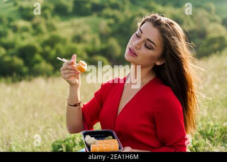 Sushi-Set und Mädchen auf grünen Hügeln Hintergrund. Lieferservice vom japanischen Restaurant. Frau mit blauen Augen in roten Kleidern Sitze und isst Sushi d Stockfoto