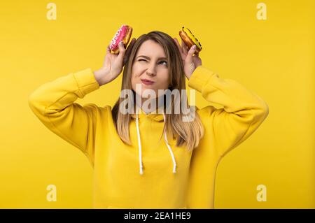 Foto von attraktiven Dame hält Donuts hinter ihrem Kopf so tun, als ob sie Hase. Trägt lässige gelbe Kapuzenpullover, isoliert gelbe Farbe Hintergrund Stockfoto