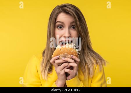 Foto von attraktiven Dame Posen in einer glücklichen Weise versucht, Burger beißen. Trägt lässige gelbe Kapuzenpullover, isoliert gelbe Farbe Hintergrund Stockfoto