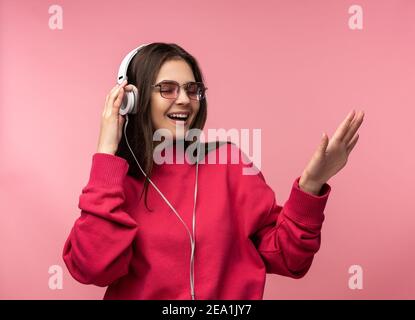 Foto von attraktiven Dame in Brille Kopfhörer Musik hören, genießt Tanzen. Trägt lässige rosa Pullover weiße Hose isoliert rosa Farbe Hintergrund Stockfoto