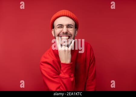 Foto von attraktiven Mann mit Bart in Brille und rote Kleidung. Männlich zeigt Rock-and-Roll-Geste und lächelt, isoliert auf rotem Hintergrund Stockfoto