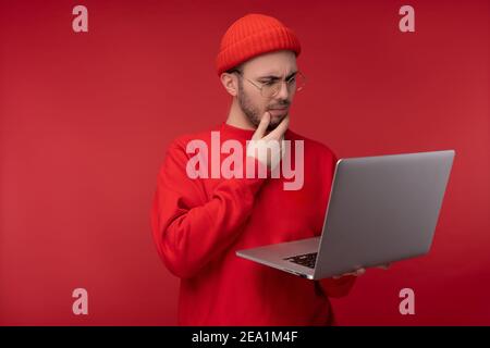 Foto von attraktiven Mann mit Bart in Brille und rote Kleidung. Serious Mann hält Laptop und Wunder, isoliert auf rotem Hintergrund Stockfoto