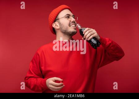 Foto von attraktiven Mann mit Bart in Brille und rote Kleidung. Glücklicher Mann hält Getränk, isoliert auf rotem Hintergrund Stockfoto