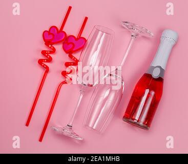 Zwei Sektgläser mit roten Plastiktrinkstrohhalmen mit Herzen und roter Sektflasche auf rosa Hintergrund. Stockfoto