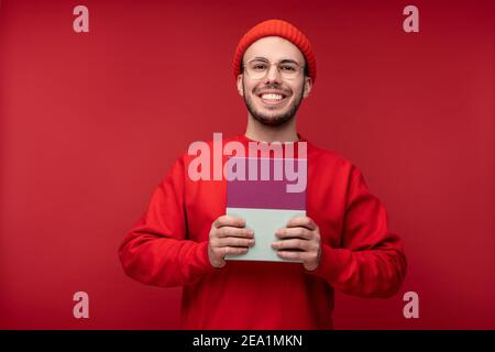 Foto von attraktiven Mann mit Bart in Brille und rote Kleidung. Männliche Griffe in Box vorhanden, isoliert auf rotem Hintergrund Stockfoto