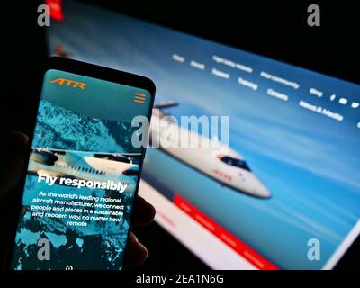Person mit Smartphone mit Website des Flugzeugherstellers Avions de Transport Régional (ATR) auf dem Bildschirm mit Hintergrund. Konzentrieren Sie sich auf die Telefonanzeige. Stockfoto