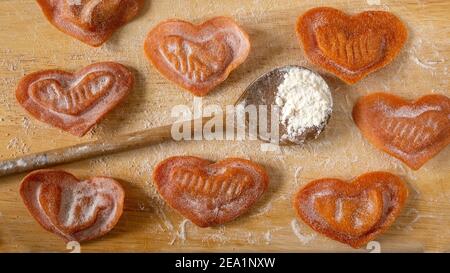 Mit Liebe gemacht, handgemachte frische herzförmige Ravioli mit Tomatenfüllung, Holzlöffel auf einem hölzernen Schneidebrett. Nahaufnahme. Stockfoto