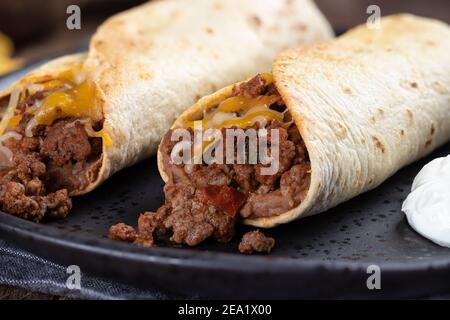 Nahaufnahme von zwei Burritos mit Hackfleisch, gebratenen Bohnen und Käse auf einem schwarzen Teller Stockfoto