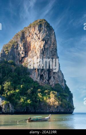 Am Railay Beach, in der Nähe von Krabi, Thailand, erstreckt sich eine riesige Felsformation ins Meer, mit einem Long-Tail-Boot auf dem Meer im Vordergrund Stockfoto