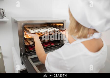 Die Arbeit des Konditoreiers. Makronen backen. Nahaufnahme der Rückansicht einer Konditorin, die Backblech mit Makronen-Keksen in den Ofen legt. Backen Stockfoto