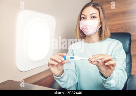 Asiatische Mädchen in einer medizinischen antiviralen Maske überprüft die Temperatur auf einem Digitalthermometer während des Fluges. Konzept eines Coronavirus covid-19 und pneumo Stockfoto