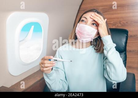 Asiatische Mädchen in einer medizinischen antiviralen Maske überprüft die Temperatur auf einem Digitalthermometer während des Fluges. Konzept eines Coronavirus covid-19 und pneumo Stockfoto