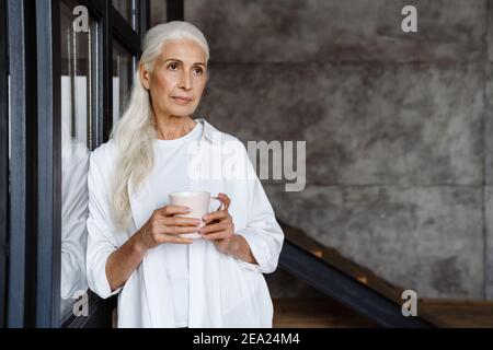 Ältere Frau trinkt Kaffee in der Nähe eines Fensters zu Hause Stockfoto