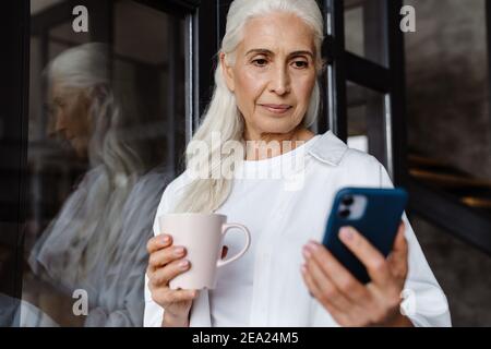 Lächelnde ältere Frau trinkt Kaffee in der Nähe eines Fensters zu Hause, mit dem Handy Stockfoto
