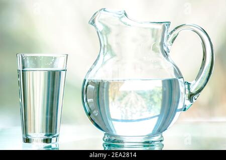 Ein klarer Krug und ein Glas gefüllt mit Wasser. Es wird direkt auf einem Glastisch geschossen, mit einem Naturhintergrund durch ein Fenster gesehen .Shallow DOF. Stockfoto