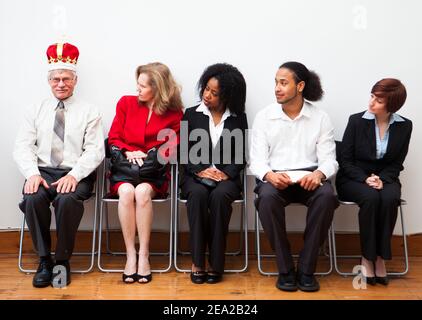 Eine Gruppe potenzieller Mitarbeiter, die auf ein Interview warten. Ein Mann trägt eine Krone, um sich von den anderen abzuheben. Humorvolles Geschäftskonzept. Stockfoto