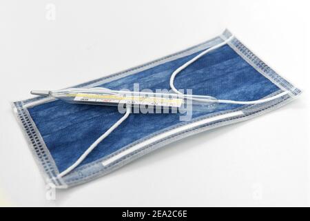 Blaue medizinische Maske und medizinisches Quecksilber-in-Glas-Thermometer auf weißem Hintergrund. Stockfoto