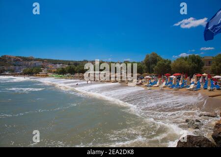 Kalyves, KRETA-GRIECHENLAND: JULI 5 2017.der Strand Kalyves.Rote Sonnenliege und Sonnenschirme, Menschen auf Hintergrund Häuser von Dorf. Stockfoto