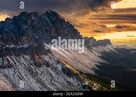 Felsen und Felsen der verschneiten Geisler Berge und dramatischer Himmel mit golden glühenden Wolken bei Sonnenuntergang im Herbst, Dolomiten, Südtirol, Italien Stockfoto