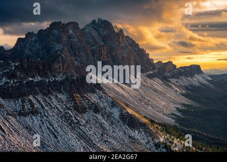 Felsen und Felsen der verschneiten Geisler Berge und dramatischer Himmel mit golden glühenden Wolken bei Sonnenuntergang im Herbst, Dolomiten, Südtirol, Italien Stockfoto