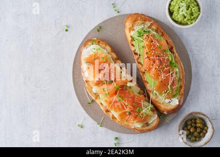 Zwei offene Sandwiches, Toast mit Lachs, Frischkäse, Avocado, Gurkenscheiben auf weißem Betontisch Stockfoto