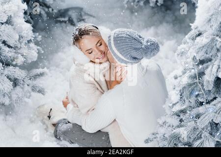 Mann küsst und umarmt seine Frau und Schnee fällt. Liebe zum neuen Jahr. Die Menschen trugen Pelzkopfhörer, Hüte, weiße Pullover. Stockfoto
