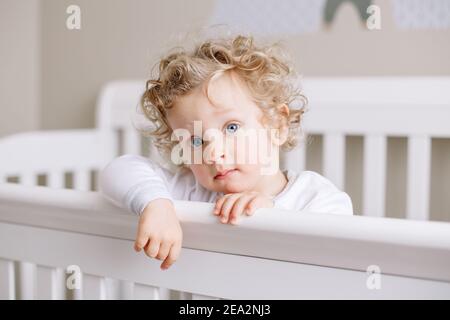 Niedliche liebenswert Baby junge Kleinkind im Kinderbett im Kinderzimmer zu Hause stehen. Neugierige charmante junge mit lockigen blonden Haaren und blauen Augen suchen Stockfoto