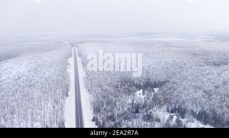 Luftaufnahme einer Straße, die durch einen verschneiten Wald führt An einem bewölkten Tag Stockfoto