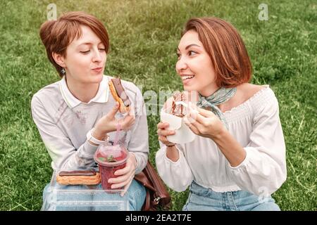 Zwei Freundinnen machen ein Picknick im Stadtpark auf grünem Gras. Street Food und Kommunikationskonzept Stockfoto