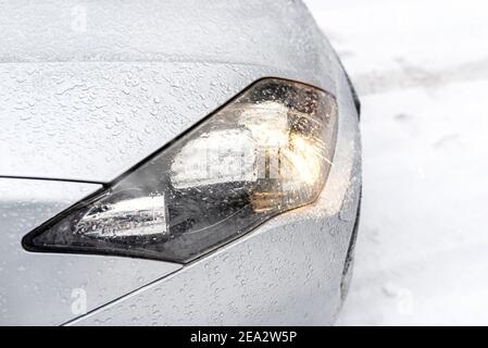 Die abgetauchten Scheinwerfer auf einem silbernen Auto stehen auf einer verschneiten Straße im Wald, sichtbaren Schnee fallen. Stockfoto