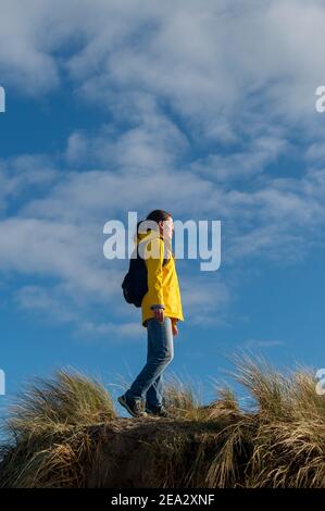 Weibliche Wanderin, die entlang hoher Sanddünen geht. Gelbe Jacke und Rucksack gegen einen blauen Himmel. Stockfoto