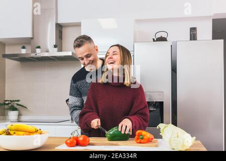 Stock Foto von einem jungen Paar lachen und kochen gesunde Lebensmittel zusammen in der Küche zu Hause. Gemüse für eine vegane Mahlzeit schneiden