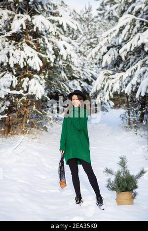 Ein junges, schönes Mädchen in einem grünen Pullover und Hut steht im Winterwald mit einem Korb mit Kiefernästen und einem Saitenbeutel mit Mandarinen. Stockfoto