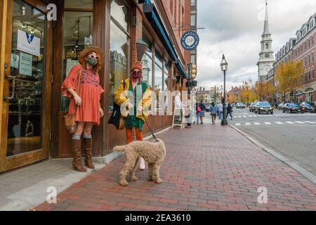 Zwei bunt gekleidete Frauen in Masken mit ihrem Hund auf der Straße der Stadt Portsmouth, NJ mit Stadtbild im Hintergrund Stockfoto