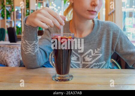 Eine junge Frau in einem grauen Pullover sitzt an einem Tisch in einem Café vor einem Glas Glühwein. Selektiver Fokus. Stockfoto