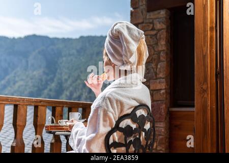 Die Frau beim Frühstück sitzt auf einem Stuhl auf dem Balkon mit Blick auf die Berge in Spanien. Mädchen im Bademantel in einem Urlaub Spa Retreat Stockfoto