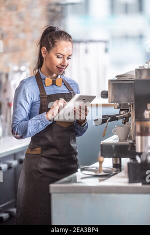 Kellnerin benutzt Tablet, während sie wartet, bis Espressos in einer Kaffeemaschine fertig sind. Stockfoto